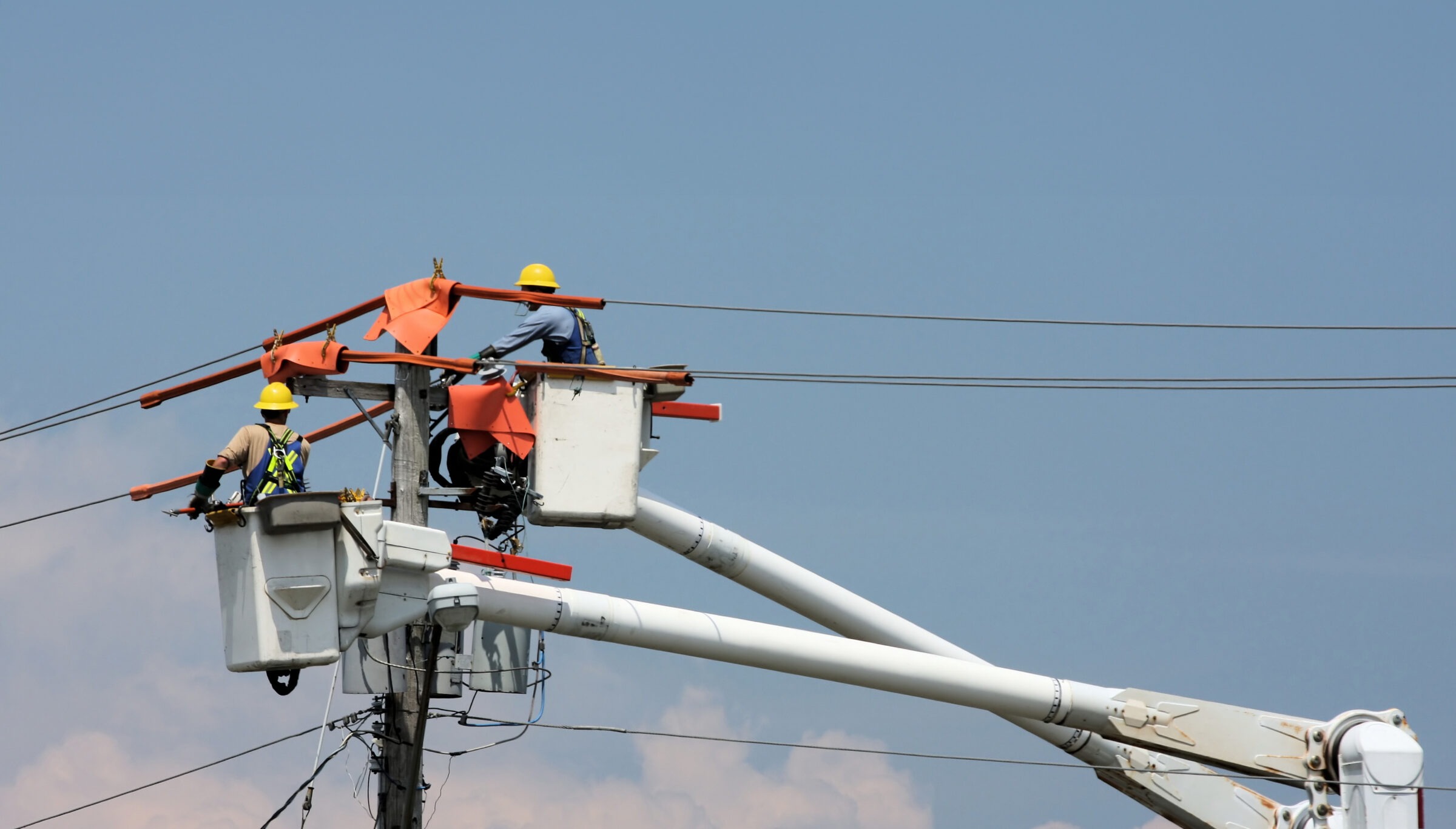Two people in safety gear are working on electrical lines from a bucket truck against a clear blue sky background. Safety cones are visible.