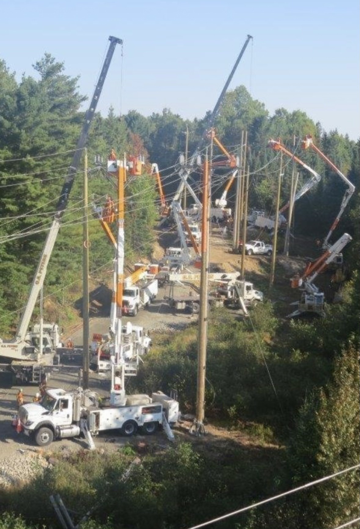 Aerial view of an outdoor industrial site with multiple utility vehicles and cranes, people working on electrical poles amidst forested area.